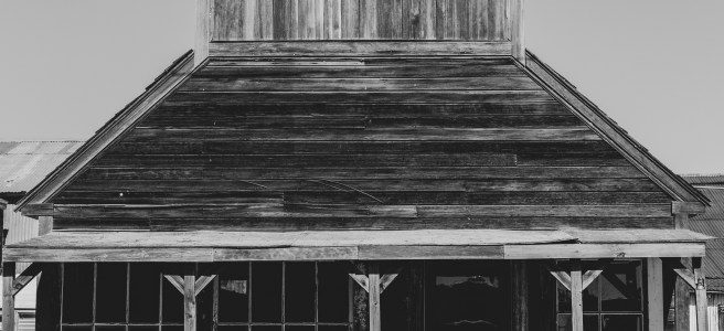 Black and white photograph of an abandoned frontier-era building with wooden walkway, benches, and distinctive peaked roof. The weathered structure features vertical plank construction and covered porch supported by wooden posts.